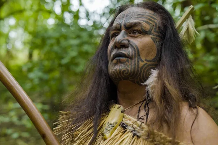 Person with traditional tattoo and costume holding a wooden staff in a forest.