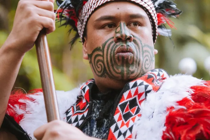 Person in traditional attire with face markings holding a staff outdoors.