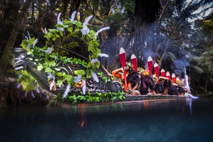 Decorated canoe with people in traditional attire, adorned with leaves and feathers on a river.
